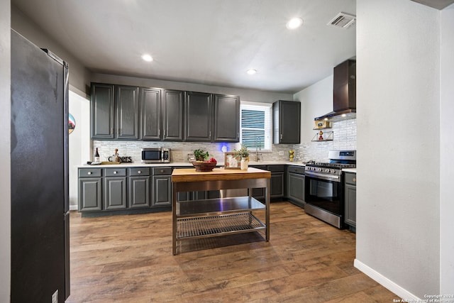 kitchen with wood-type flooring, appliances with stainless steel finishes, wall chimney range hood, and backsplash