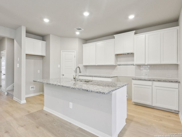 kitchen with white cabinets, sink, light wood-type flooring, light stone counters, and a center island with sink