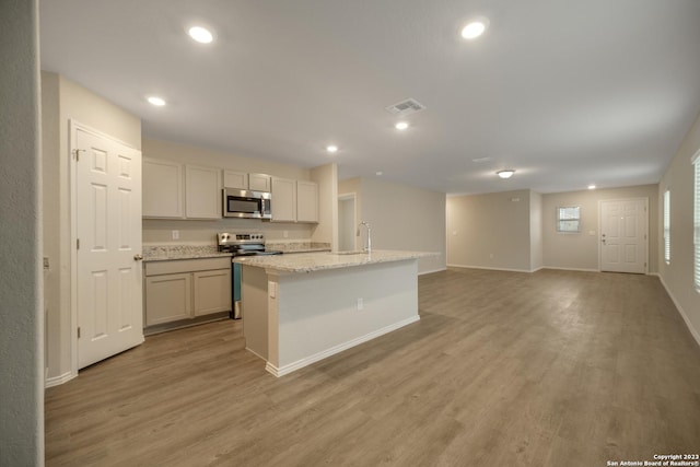 kitchen with appliances with stainless steel finishes, sink, light wood-type flooring, light stone counters, and a kitchen island with sink