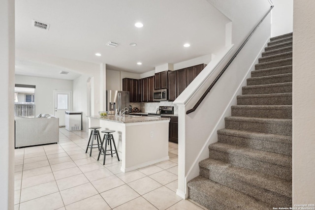 kitchen featuring light tile patterned floors, dark brown cabinetry, an island with sink, stainless steel appliances, and a breakfast bar area