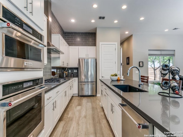 kitchen featuring appliances with stainless steel finishes, wall chimney exhaust hood, white cabinetry, sink, and light hardwood / wood-style flooring