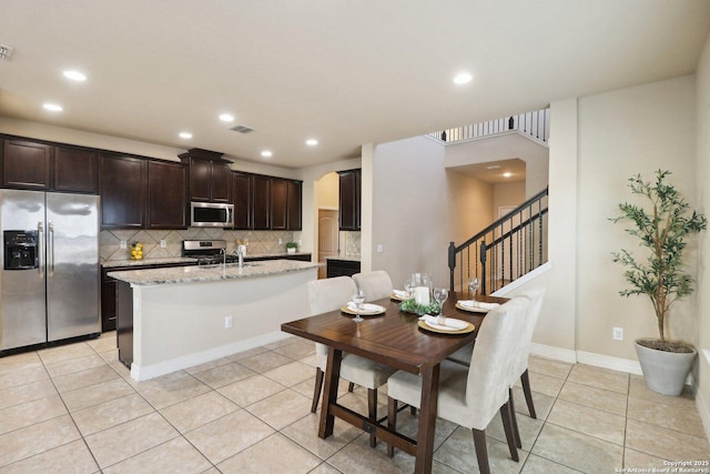 kitchen featuring light stone countertops, appliances with stainless steel finishes, a kitchen island with sink, decorative backsplash, and light tile patterned flooring