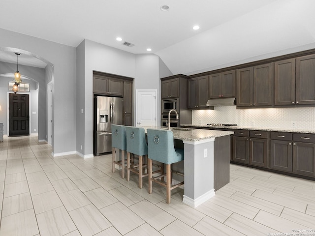 kitchen with dark brown cabinets, appliances with stainless steel finishes, backsplash, a kitchen island with sink, and lofted ceiling