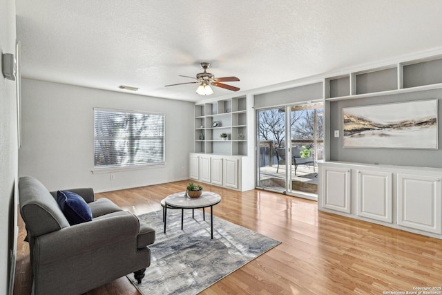 living room featuring built in shelves, ceiling fan, light hardwood / wood-style flooring, and a textured ceiling