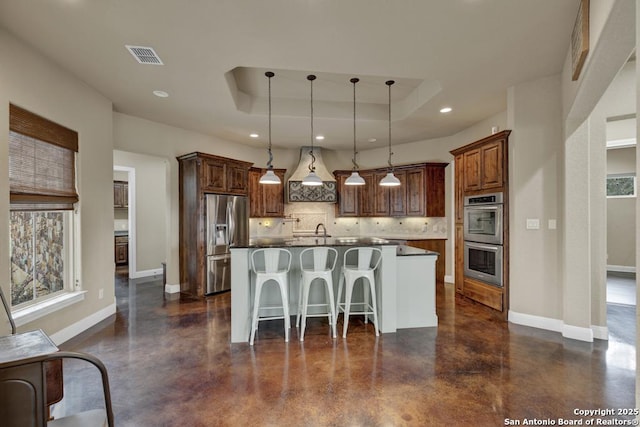 kitchen featuring tasteful backsplash, hanging light fixtures, appliances with stainless steel finishes, a tray ceiling, and an island with sink