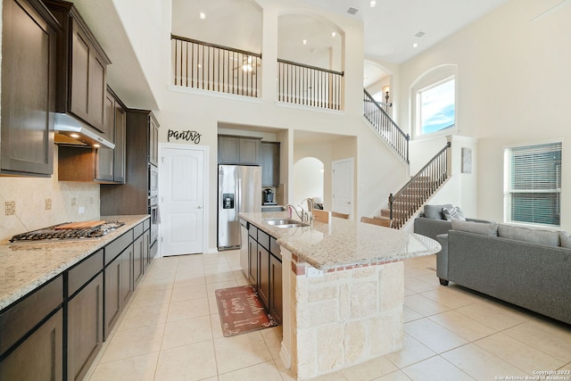 kitchen with appliances with stainless steel finishes, sink, a towering ceiling, light stone countertops, and a kitchen island with sink