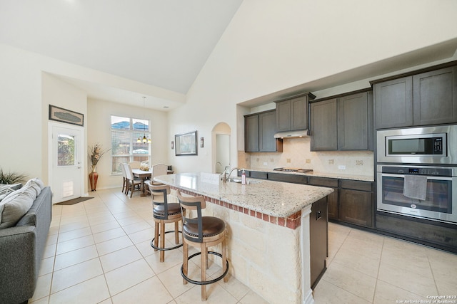 kitchen with appliances with stainless steel finishes, a kitchen breakfast bar, dark brown cabinets, light stone counters, and a center island with sink