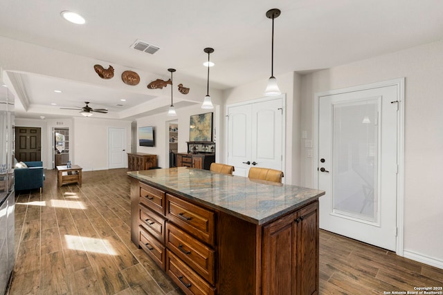 kitchen with decorative light fixtures, a tray ceiling, a kitchen island, ceiling fan, and dark stone counters