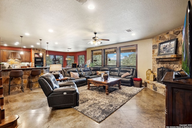 living room featuring ceiling fan, a textured ceiling, a wealth of natural light, and a stone fireplace