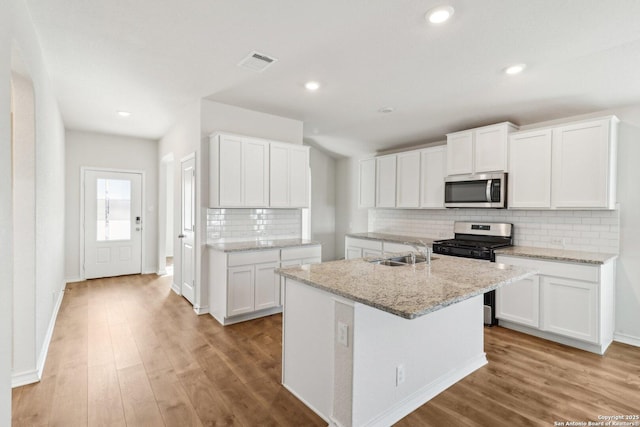 kitchen with light stone countertops, appliances with stainless steel finishes, white cabinetry, light wood-type flooring, and a center island with sink