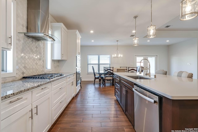 kitchen with appliances with stainless steel finishes, white cabinetry, wall chimney range hood, sink, and a kitchen island with sink