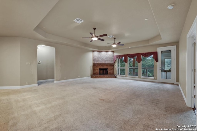 unfurnished living room featuring a raised ceiling, a brick fireplace, light colored carpet, and ceiling fan