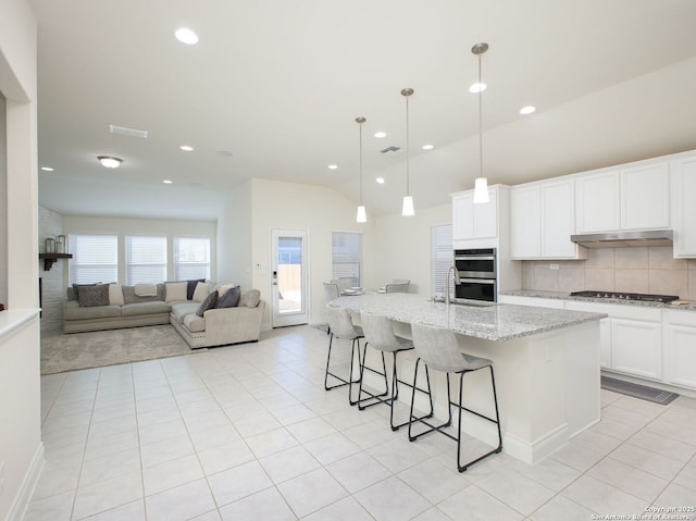 kitchen with white cabinetry, light stone counters, a center island with sink, decorative light fixtures, and vaulted ceiling