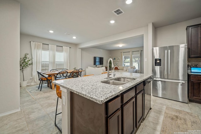 kitchen with dark brown cabinetry, sink, stainless steel appliances, light stone countertops, and a kitchen island with sink