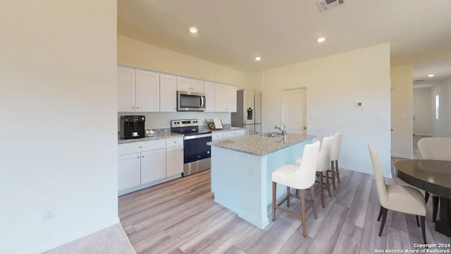 kitchen featuring appliances with stainless steel finishes, an island with sink, white cabinets, light stone counters, and light wood-type flooring