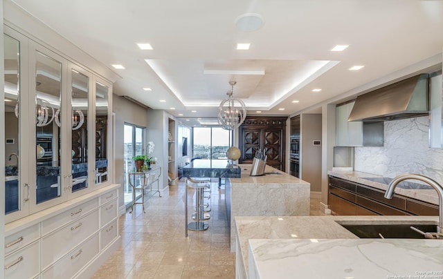 kitchen with decorative light fixtures, a breakfast bar area, white cabinets, a tray ceiling, and wall chimney exhaust hood