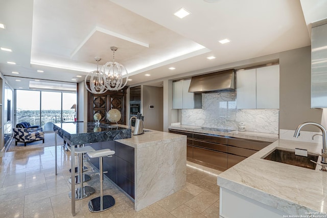 kitchen featuring sink, a kitchen island with sink, wall chimney range hood, and a tray ceiling