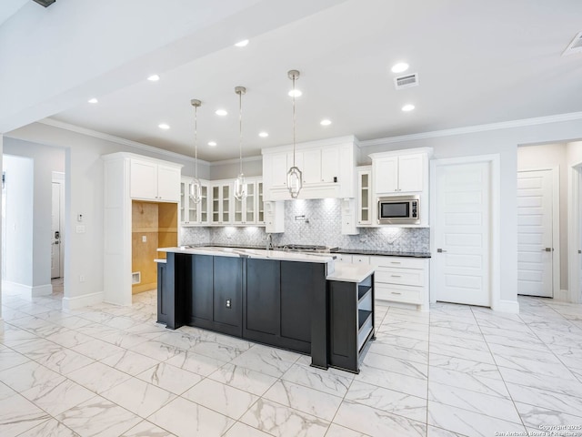 kitchen featuring a kitchen island with sink, hanging light fixtures, white cabinetry, stainless steel microwave, and ornamental molding