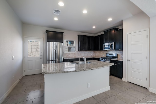 kitchen with appliances with stainless steel finishes, sink, a kitchen island with sink, light tile patterned floors, and light stone counters