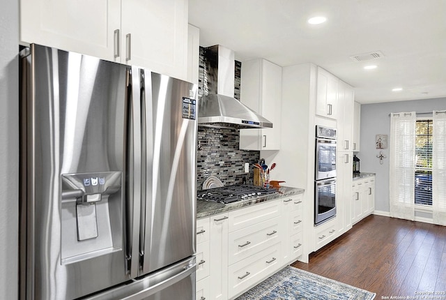 kitchen featuring wall chimney exhaust hood, dark stone countertops, backsplash, appliances with stainless steel finishes, and white cabinets