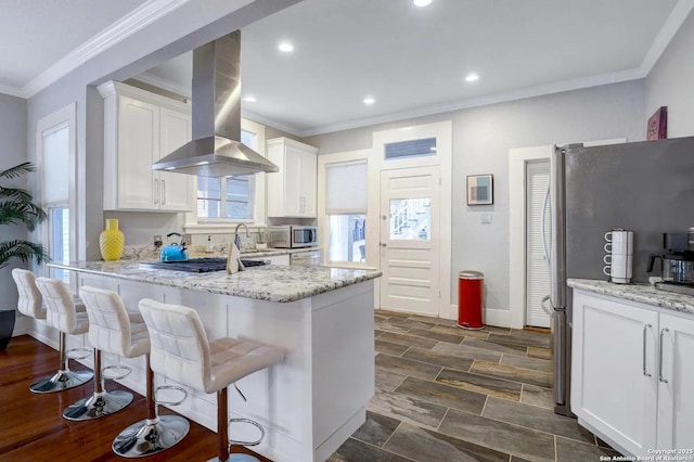 kitchen with a breakfast bar area, appliances with stainless steel finishes, white cabinetry, light stone counters, and island range hood