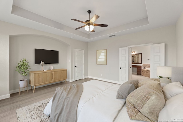 bedroom featuring ceiling fan, connected bathroom, a raised ceiling, and light hardwood / wood-style floors