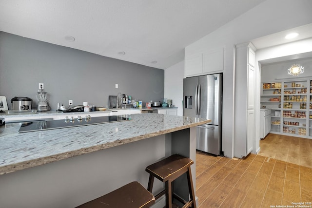 kitchen featuring white cabinetry, a breakfast bar area, black electric stovetop, stainless steel fridge with ice dispenser, and light stone countertops