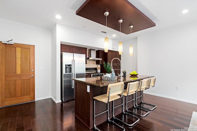 kitchen with pendant lighting, range, a kitchen island with sink, stainless steel refrigerator with ice dispenser, and wall chimney exhaust hood