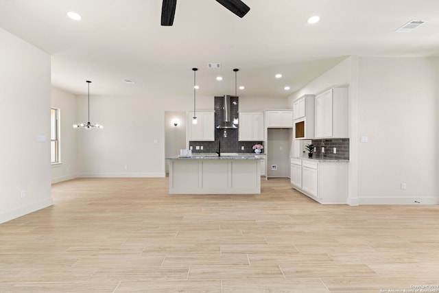 kitchen with white cabinets, decorative light fixtures, a kitchen island with sink, and wall chimney range hood