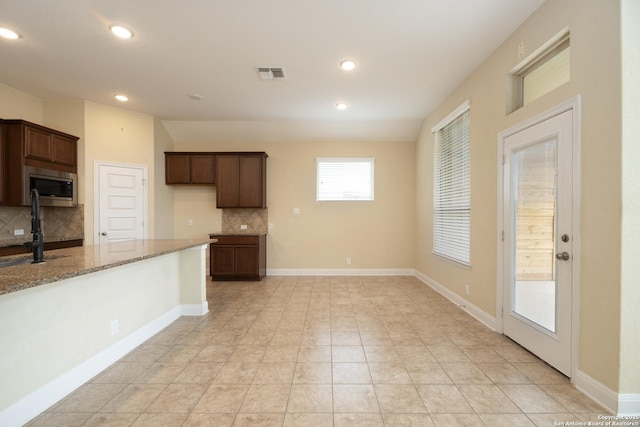 kitchen featuring light stone counters, dark brown cabinets, light tile patterned floors, stainless steel microwave, and decorative backsplash