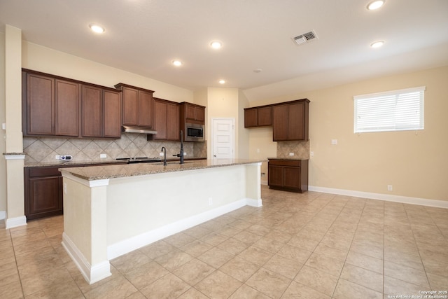 kitchen with stainless steel microwave, tasteful backsplash, a kitchen island with sink, light tile patterned floors, and light stone counters