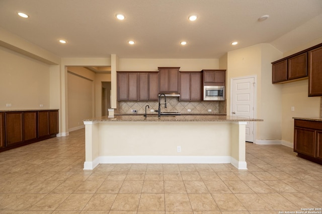 kitchen featuring sink, a kitchen island with sink, stainless steel microwave, light stone counters, and tasteful backsplash