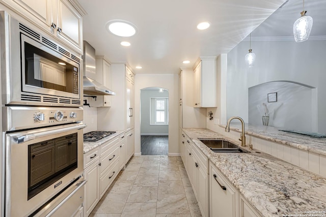 kitchen featuring wall chimney exhaust hood, sink, light stone counters, hanging light fixtures, and stainless steel appliances