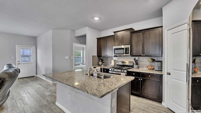 kitchen featuring sink, backsplash, stainless steel appliances, light stone countertops, and a center island with sink