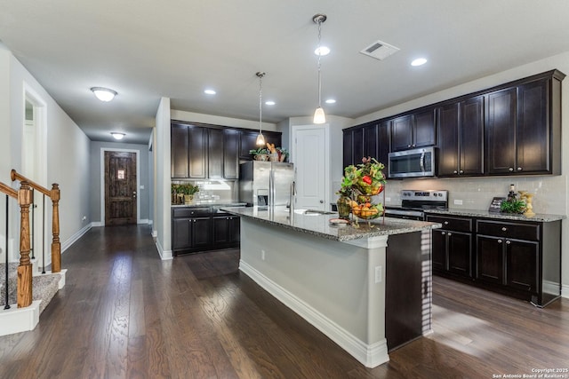 kitchen featuring light stone counters, hanging light fixtures, a center island with sink, appliances with stainless steel finishes, and dark hardwood / wood-style flooring