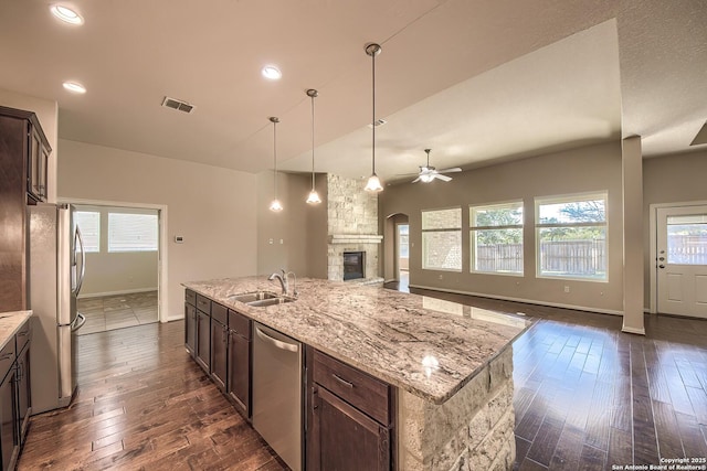 kitchen with an island with sink, sink, dark brown cabinetry, stainless steel appliances, and light stone countertops