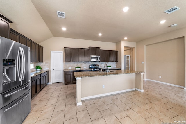 kitchen with lofted ceiling, appliances with stainless steel finishes, light stone countertops, an island with sink, and decorative backsplash