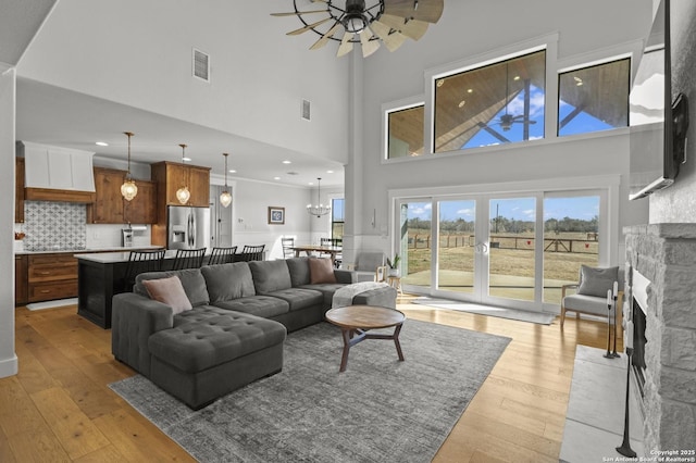 living room with french doors, a stone fireplace, light hardwood / wood-style flooring, ceiling fan with notable chandelier, and a high ceiling