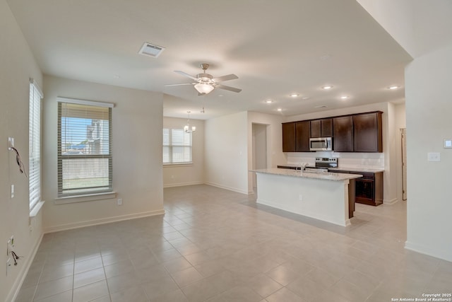 kitchen featuring ceiling fan with notable chandelier, stainless steel appliances, light stone countertops, dark brown cabinets, and a center island with sink