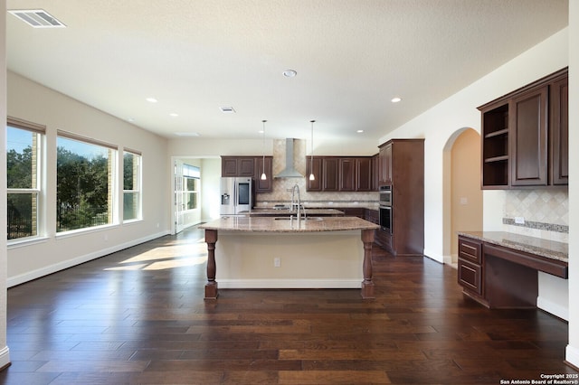 kitchen with appliances with stainless steel finishes, decorative light fixtures, wall chimney range hood, light stone countertops, and a center island with sink