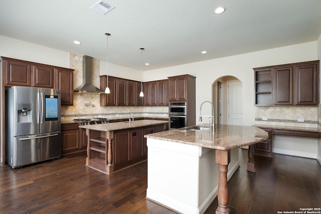 kitchen with sink, a center island with sink, pendant lighting, stainless steel appliances, and wall chimney range hood