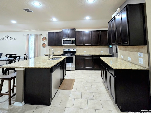 kitchen with sink, light stone counters, a center island with sink, stainless steel appliances, and decorative backsplash