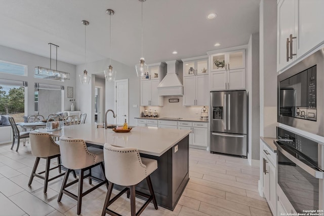 kitchen featuring white cabinets, a kitchen bar, a kitchen island with sink, stainless steel appliances, and custom range hood