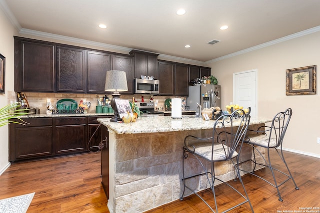 kitchen with crown molding, dark wood-type flooring, appliances with stainless steel finishes, a kitchen breakfast bar, and a center island with sink