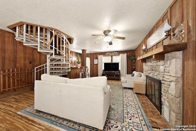 living room with ceiling fan, a stone fireplace, wooden walls, and a textured ceiling