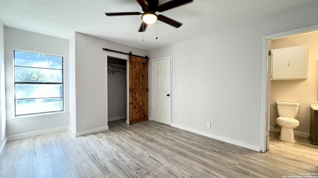 unfurnished bedroom with connected bathroom, a closet, ceiling fan, a barn door, and light hardwood / wood-style floors