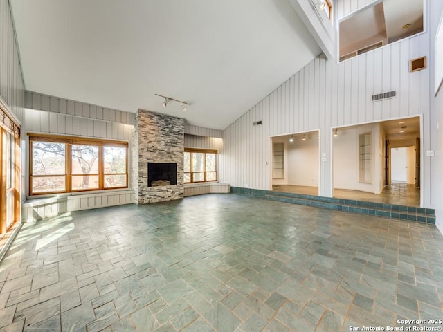 unfurnished living room featuring rail lighting, a stone fireplace, high vaulted ceiling, and wood walls