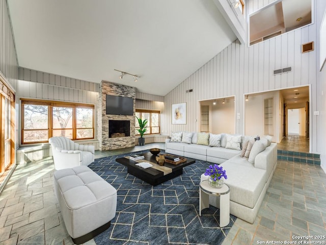 living room featuring rail lighting, a stone fireplace, high vaulted ceiling, and wood walls