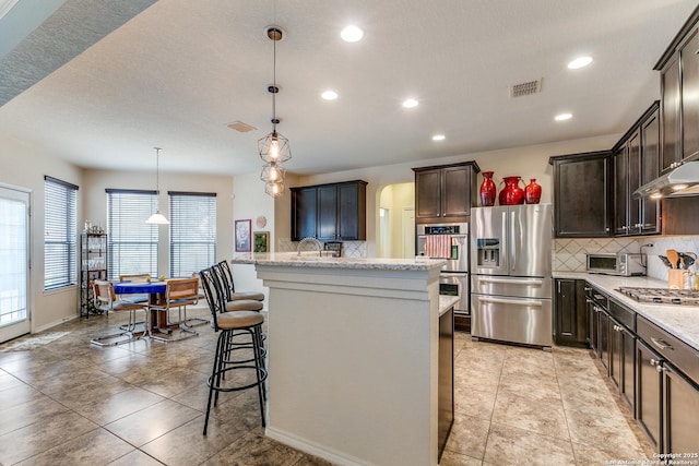 kitchen featuring appliances with stainless steel finishes, backsplash, hanging light fixtures, dark brown cabinets, and a center island with sink