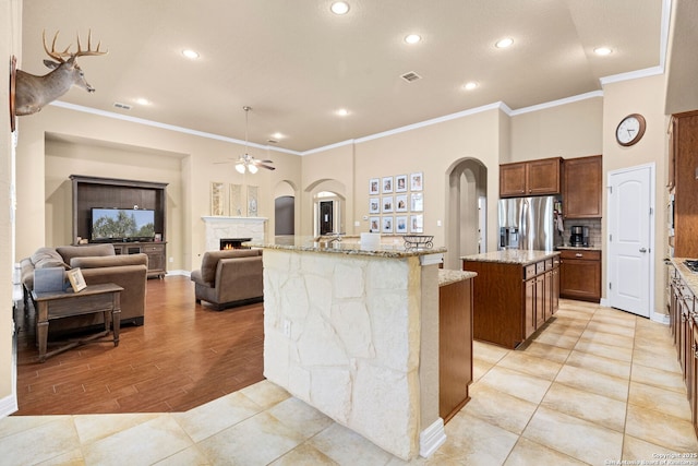 kitchen featuring stainless steel fridge, ceiling fan, light stone counters, crown molding, and a center island with sink